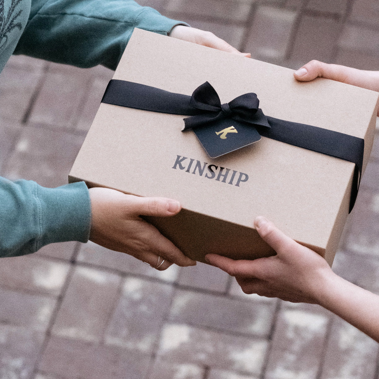 two sets of hands handing off a Kinship holiday gift box with black ribbon and a tag