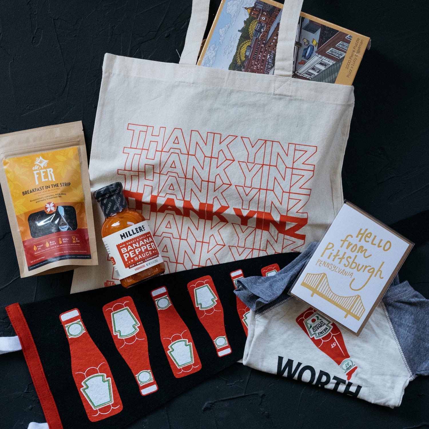 Kinship flatlay showing Pittsburgh-themed items including a Heinz ketchup pennant, a Heinz ketchup baby onesie, a Meshwork Press card, Millers hot sauce, De Fer Breakfast in the Strip tea and a Thank Yinz tote